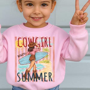 Może przedstawiać: R&oacute;żowa bluza z napisem "COWGIRL SUMMER" i grafiką kowbojki z deską surfingową. Projekt zawiera słońce, palmy i słowo "HOWDY!"