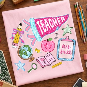 May include: A light pink t-shirt featuring a colorful graphic print of school supplies including a pencil with a pink bow, a globe, a magnifying glass, an open book, a ruler, a star, a smiley apple, and a clipboard labeled Mrs. Smith.