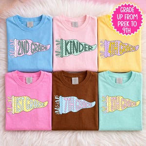 May include: Six colorful t-shirts with grade level designs. The shirts are light blue, pink, yellow, brown, and mint green. Each shirt features a pennant design with the grade level printed on it, from 1st to 9th grade.