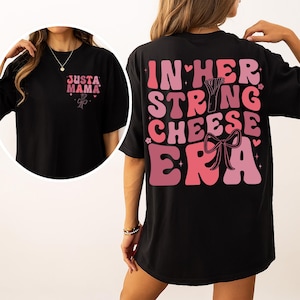 Könnte beinhalten: Schwarzes T-Shirt mit rosa Text und Grafiken. Auf der Vorderseite des Shirts steht "Just a Mama" mit einer Schleifengrafik. Auf der Rückseite des Shirts steht "In Her String Cheese Era" in rosa Retro-Schrift mit einer Schleifengrafik.