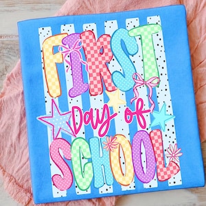 May include: A blue t-shirt with the words "FIRST DAY OF SCHOOL" in colorful, patterned letters. The design includes stars, bows, and floral accents. The background has white vertical stripes with black polka dots.