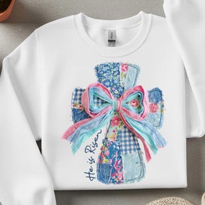 Pode incluir: Camisola branca com um design de cruz de retalhos com padrões florais, ganga e xadrez. Um grande laço de fita rosa e azul adorna a cruz. As palavras "He is Risen" estão escritas na camisola.