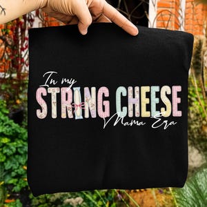 May include: Black t-shirt with the text "In my STRING CHEESE Mama Era" in a colorful, patterned font. The words are in a mix of pastel colors and floral patterns.