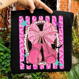 May include: A black t-shirt featuring a graphic of two pink boxing gloves tied together with a large pink bow patterned with breast cancer awareness ribbons. The design includes vertical pink striped bands with bat silhouettes, teal stars, and a scalloped border.