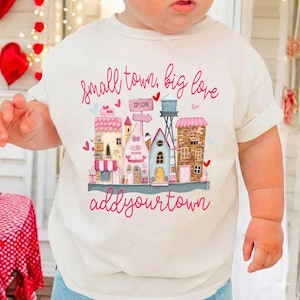 Puede incluir: Camiseta color crema con una ilustración de pueblo caprichosa. Edificios rosas y azules, corazones y el texto "small town, big love" y "add your town" en escritura rosa. Un diseño encantador.