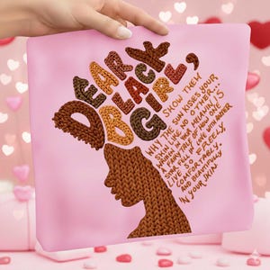May include: A pink pillow with the words "Dear Black Girl" in a knitted font, with a silhouette of a woman's face. The text "Why the sun kisses your skin like no other..." is also on the pillow.