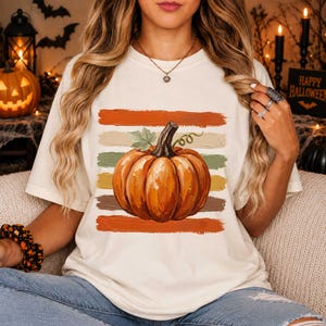 May include: A cream-colored t-shirt featuring a central graphic of a large orange pumpkin with a green stem and leaf. The pumpkin is layered over five horizontal brushstroke stripes in shades of orange, tan, olive green, and brown.