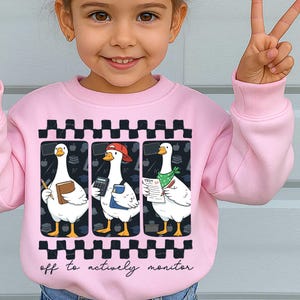 Peut inclure: Sweat-shirt rose avec un motif de trois oies de dessin anim&eacute;, chacune tenant des fournitures scolaires. Les oies sont dans des cadres noirs avec une bordure &agrave; carreaux. Le texte "off to actively monitor" est en dessous.