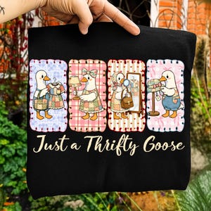Puede incluir: Camiseta negra con un gráfico caprichoso de un ganso con un vestido de retazos, sosteniendo un vestido, una lámpara, un espejo y sombreros. El texto "Just a Thrifty Goose" está debajo del gráfico. El diseño está enmarcado con un borde rosa y azul.