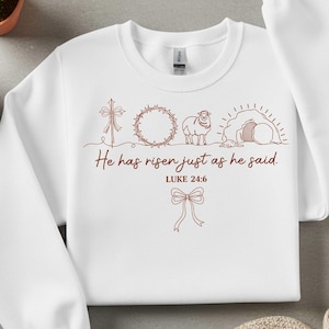 May include: White sweatshirt with a brown graphic design featuring a cross, crown of thorns, lamb, and empty tomb. The text "He has risen just as he said. Luke 24:6" is printed below the images, with a bow at the bottom.