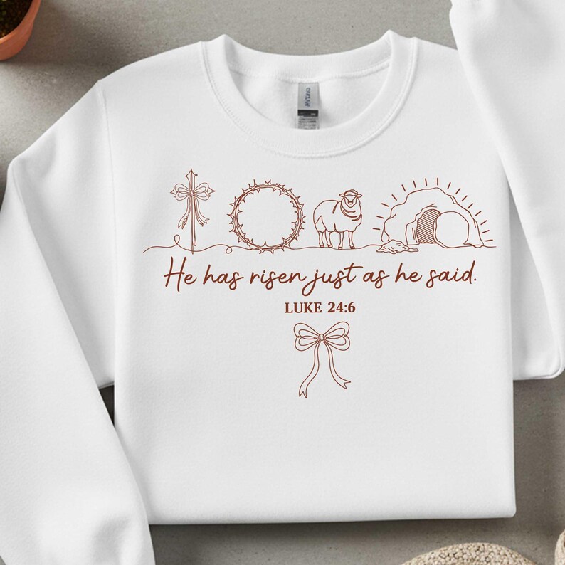 May include: White sweatshirt with a brown graphic design featuring a cross, crown of thorns, lamb, and empty tomb. The text "He has risen just as he said. Luke 24:6" is printed below the images, with a bow at the bottom.