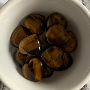 May include: A white bowl filled with tiger's eye heart-shaped stones. The stones are a mix of brown and gold colours.
