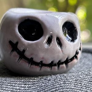 May include: A ceramic planter shaped like Jack Skellington's head from The Nightmare Before Christmas. The planter is white with black eyes, a stitched mouth, and pink accents. The planter is sitting on a gray surface.