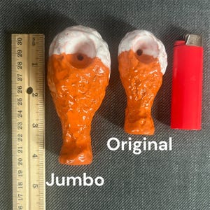 May include: Two ceramic chicken leg pipes, one labeled "Jumbo" and the other "Original". The pipes are orange and white with a hole in the top for smoking.