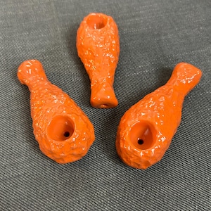 May include: Three orange ceramic pipes shaped like fried chicken drumsticks. Each pipe has a textured surface and a bowl opening. The pipes are arranged on a gray denim surface.