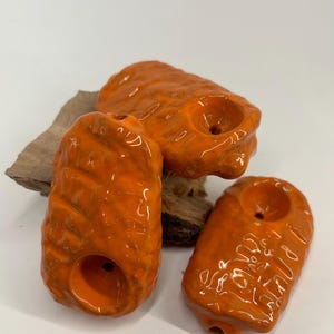 May include: Three orange ceramic pipes with a rough, textured surface. Each pipe has a round hole for smoking.