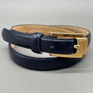 May include: A navy blue leather belt with a gold-colored buckle. The belt has a rectangular buckle with a V-shaped design. The belt is approximately 1 inch wide.