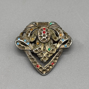 Puede incluir: Broche vintage plateado con diseño floral y lazo. El broche está adornado con pedrería de colores rojo, azul, verde y amarillo. El broche tiene un diseño calado y una forma geométrica.