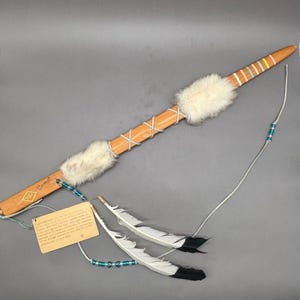 May include: A handcrafted wooden ceremonial staff with white fur accents and a decorative feather and bead adornment. The staff features a light brown finish, with colorful stripes near the tip. The staff is approximately 30 inches long.