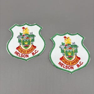 May include: Two white embroidered patches with a green border. Each patch features a shield design with a crest, the words "FORGE AHEAD" and "NELSON B.C." in red. The crest includes a crown, lion, and foliage.