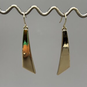 Puede incluir: Un par de pendientes colgantes de tono dorado con una forma triangular alargada, lisa y pulida. Cada pendiente cuelga de un sencillo gancho de alambre curvado, mostrando una superficie metálica reflectante que se estrecha hacia abajo.