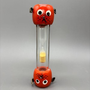 May include: A vertical hourglass featuring two orange wooden ends shaped like stylized animal heads with black painted eyes, noses, and whiskers. The central glass cylinder contains yellow sand, held in place by thin metal support rods.