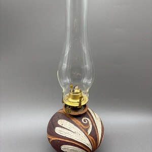 May include: An oil lamp with a clear glass chimney and a brass burner mechanism. The base is a round, dark brown ceramic vessel featuring carved, light-colored leaf-like patterns that wrap around the exterior of the rounded body.