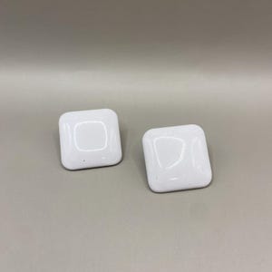 May include: Two white, square cabinet knobs with rounded edges. Each knob has a recessed square in the center and a speckled finish. The knobs are set against a neutral gray background.