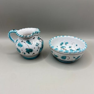 以下が含まれることがあります： 青と緑の花柄が描かれた白い陶器のクリーマーとボウル。クリーマーはハンドルと注ぎ口が付いています。ボウルは丸く、縁がギザギザになっています。