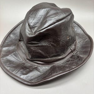 May include: A dark brown leather hat featuring a creased crown and a wide, flexible brim. The hat is constructed from multiple stitched leather panels and includes small metal ventilation eyelets on the sides of the crown for airflow.