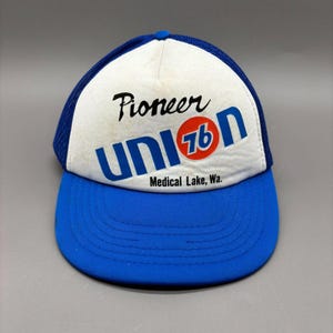 Pode incluir: Boné de caminhoneiro azul e branco com o texto "Pioneer Union 76 Medical Lake, Wa.". O boné tem uma aba azul e um painel frontal branco com o logotipo da Union 76. A parte de trás do boné é de malha azul.