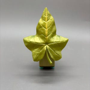 Puede incluir: Un objeto decorativo en forma de hoja de color dorado. La hoja tiene vetas detalladas y una superficie ligeramente texturizada. La base es de forma rectangular con dos pequeños acentos circulares de color turquesa. El objeto está sobre un fondo gris.