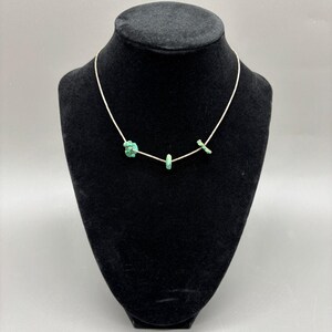 May include: A delicate necklace featuring a thin silver chain with three turquoise-colored stone accents. The stones are irregularly shaped, adding a unique, natural aesthetic. The necklace is displayed on a black mannequin, highlighting the jewelry's design.