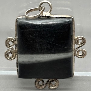 May include: A square pendant featuring a polished black stone with a horizontal white band. The stone is set in a silver-toned metal frame with decorative scrollwork accents on the left, right, and bottom edges, and a circular bail at the top.