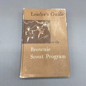May include: A brown book with the title "Leader's Guide to the Brownie Scout Program". The cover features an illustration of three girls in a forest setting.