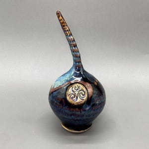 May include: A handcrafted ceramic vase with a bulbous base and a long, curved spout. The vase features a dark blue glaze with brown and turquoise accents. A gold-colored Celtic knot design is centered on the front.