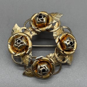 Pode incluir: Broche vintage em metal dourado com design circular. O broche apresenta cinco rosas detalhadas com centros prateados e folhas. O alfinete está sobre um fundo cinza.