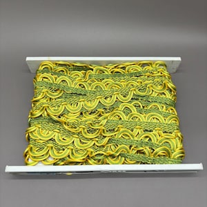 May include: A spool of decorative gimp trim featuring a repeating scalloped pattern in vibrant yellow and lime green. The trim is wound tightly around a white rectangular cardboard holder, showcasing a textured weave and a glossy finish.
