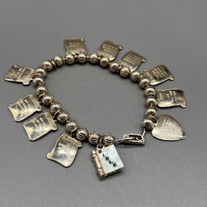 May include: A gold-toned bracelet featuring scroll-shaped charms with engraved text and a heart-shaped charm. The bracelet also includes a small book charm with the word "BIBLE" and the numbers "1872". The bracelet is strung with gold-colored beads.