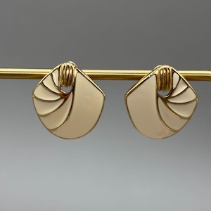 May include: A pair of vintage gold-tone earrings with a cream-colored enamel design. The earrings have a fan-like shape with gold accents and a post-back closure. The earrings are displayed on a gold bar.