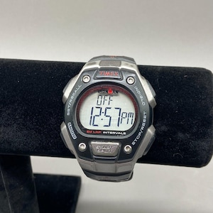 Puede incluir: Un reloj digital Timex Ironman con caja negra y plateada. La esfera muestra "OFF" y la hora "12:57 pm". Incluye botones "Set/Recall", "Mode", "Start/Split" y "Stop/Reset". El reloj está sobre una superficie negra.
