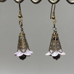 May include: A pair of dangle earrings with a floral design. Each earring features a bronze-colored filigree cone with a white and pink flower-shaped base and a dark, round bead at the bottom. The earrings hang from bronze-colored hooks.
