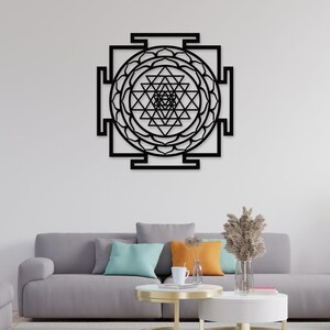 Puede incluir: Arte de pared de metal negro con un dise&ntilde;o geom&eacute;trico de un Sri Yantra, un s&iacute;mbolo sagrado en el hinduismo. El arte se muestra en una pared blanca sobre un sof&aacute; gris con almohadas.