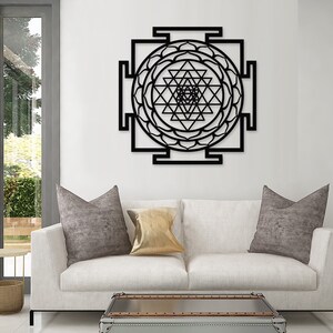 Puede incluir: Arte de pared de metal negro con un dise&ntilde;o geom&eacute;trico de un Sri Yantra, un s&iacute;mbolo sagrado en el hinduismo. El arte se muestra en una pared blanca sobre un sof&aacute; blanco con almohadas grises y doradas.