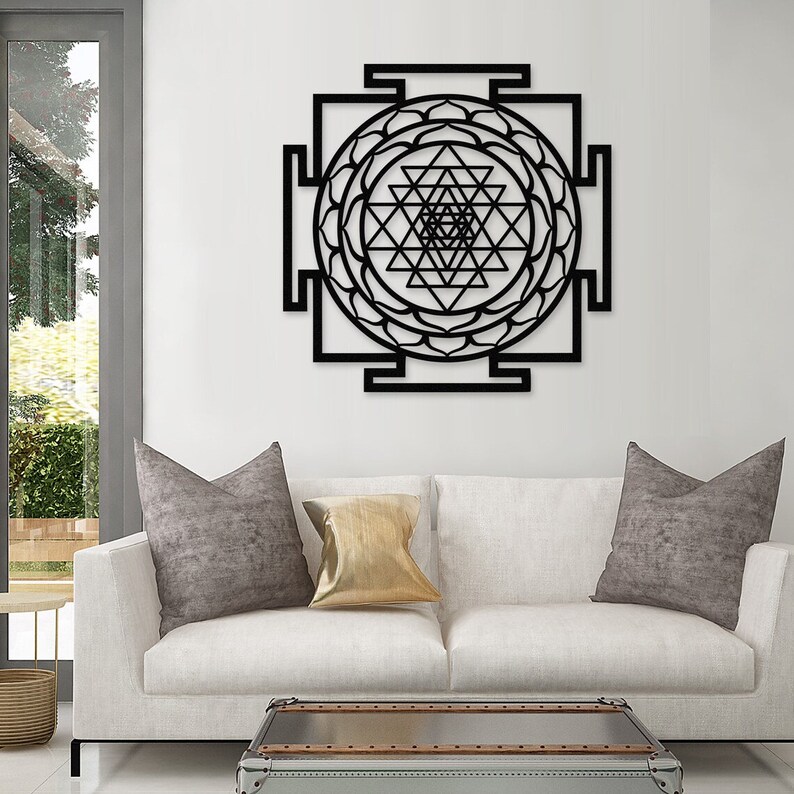 Puede incluir: Arte de pared de metal negro con un dise&ntilde;o geom&eacute;trico de un Sri Yantra, un s&iacute;mbolo sagrado en el hinduismo. El arte se muestra en una pared blanca sobre un sof&aacute; blanco con almohadas grises y doradas.