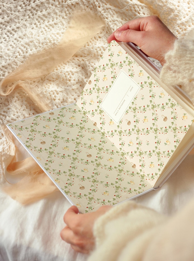 May include: An open white journal with a floral pattern on the inside pages. The pattern features green vines, red berries, and small yellow baskets. The text "Daily Notebook 2023" is printed on the back cover.