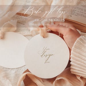 May include: Two round, white gift tags with a delicate, gold script that reads "Your design here". The tags are on a background of soft, peach-colored fabric and ribbon.