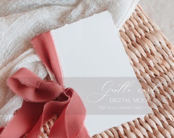 May include: A white card with a torn edge, tied with a dusty rose ribbon, sits on a woven wicker surface. The card features the text "Gentle card DIGITAL MOCKUP PSD WITH SMART OBJECT .JPEG". A white cloth is in the background.