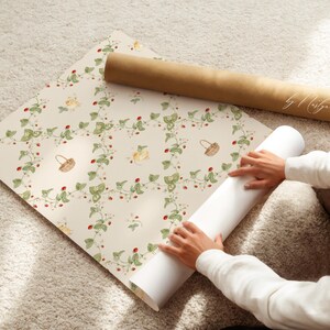 May include: A roll of wallpaper with a repeating pattern of green vines, red strawberries, and small baskets. The wallpaper is being unrolled by a person's hands.