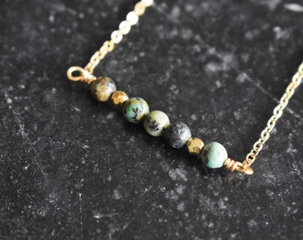 May include: A delicate gold necklace with a row of small, round beads in shades of green, grey, and brown. The beads are interspersed with gold-coloured, faceted beads. The necklace has a simple, elegant design.